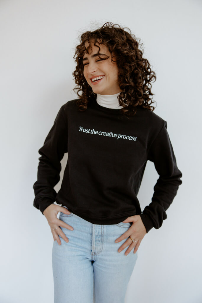 woman wearing a black sweatshirt, white turtleneck, and jeans, smiling. the sweatshirt reads "trust the creative process" in white