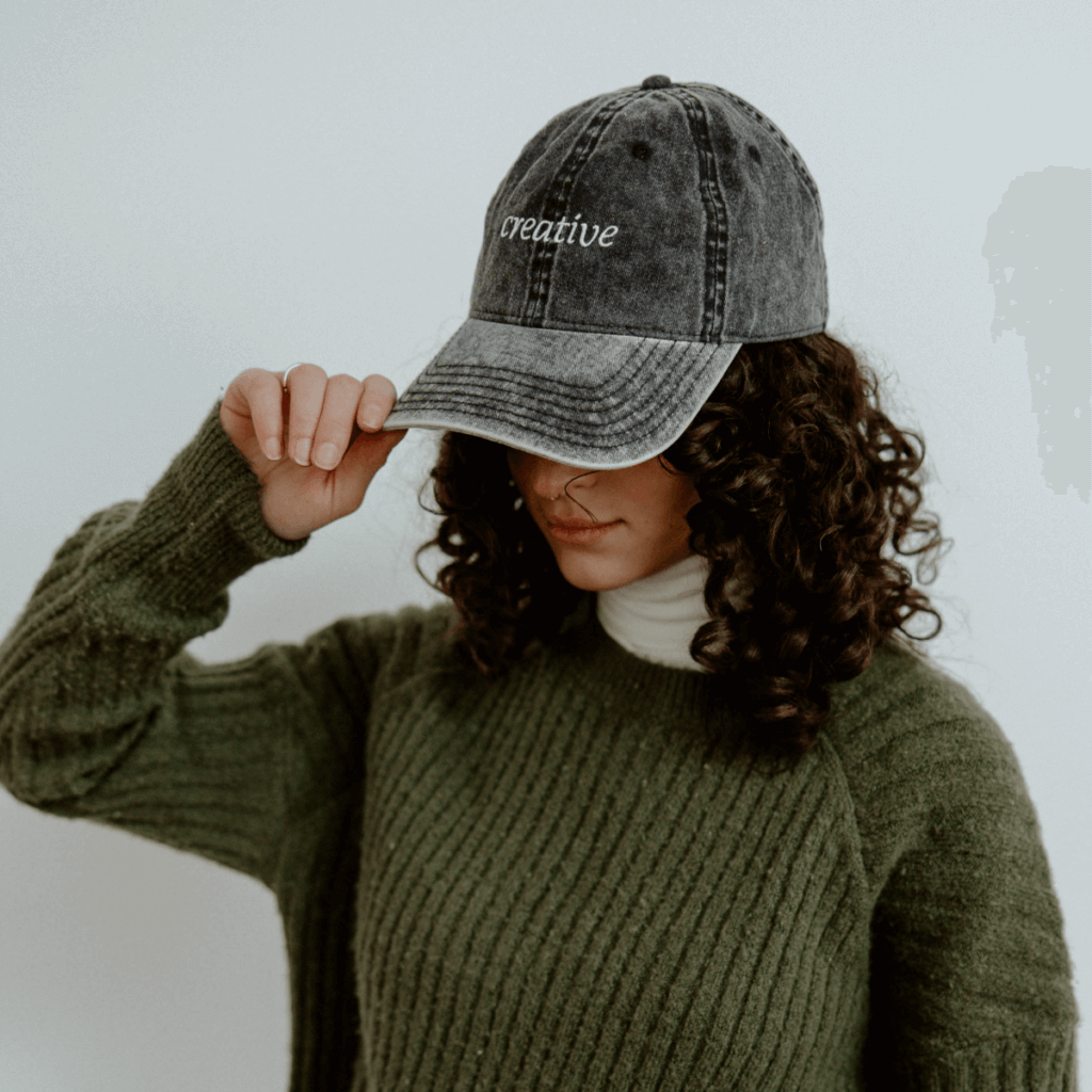 woman with curly hair wearing a black baseball cap that reads "creative", a green sweater, and a white turtleneck